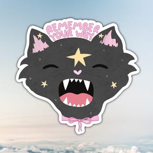 Puede incluir: Una pegatina con la cabeza de un gato negro con la boca abierta y dientes visibles. El gato tiene orejas rosas, un lazo rosa y una estrella amarilla en la frente. El texto "REMEMBER YOUR WHY" está sobre la cabeza del gato.