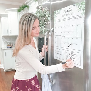 Può includere: Un magnete calendario bianco e verde su un frigorifero in acciaio inossidabile. Il calendario è datato novembre 2019 e ha una sezione note. Una donna scrive sul calendario con un pennarello nero.