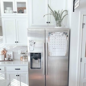 May include: A stainless steel Samsung refrigerator with a calendar and a memo board attached. The calendar displays the month of June 2021. The kitchen features white cabinets, a white countertop, and a vase of eucalyptus.