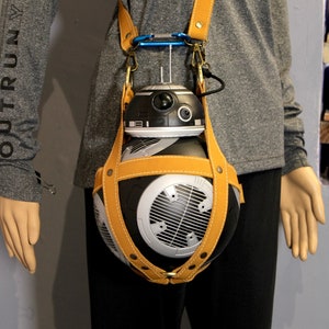 May include: A black and silver robot toy with a brown leather carrying case and a strap. The robot has a round body with a dome-shaped head and a single eye.