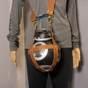 May include: A black and silver BB-8 droid-shaped bag with brown leather straps and gold-colored hardware. The bag is suspended from a brown leather shoulder strap with a blue carabiner clip. The bag is worn by a mannequin.