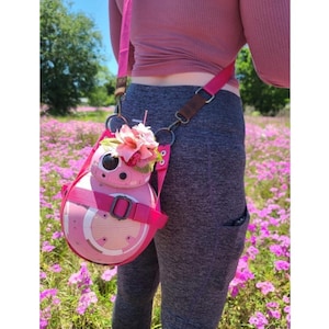 May include: A pink and white BB-8 droid-shaped crossbody bag with a floral embellishment. The bag is worn by a person in a pink shirt and gray pants.