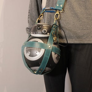 May include: A teal blue leather crossbody bag with a black and silver droid figure inside. The bag has a strap that goes over the shoulder.