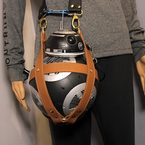 May include: A brown leather strap with metal accents holds a black and silver BB-8 droid replica. The strap is worn over the shoulder of a person wearing a gray shirt.