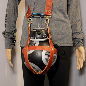May include: A brown leather strap with a black and silver robot attached. The robot has a round body with a black and white design. The strap is worn over the shoulder of a person.