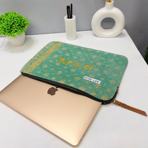 May include: A teal laptop sleeve with a floral pattern and the word "HOPE" embroidered in gold. The sleeve has a black zipper and a brown leather pull. The laptop is gold.