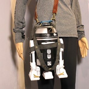 May include: A black and white robot character with a white body and black head and legs is attached to a brown leather strap. The strap is worn over the shoulder of a person wearing a gray shirt.