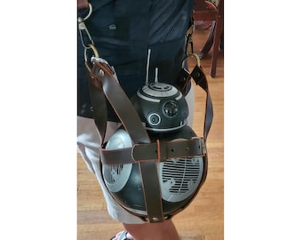 May include: A brown leather strap with metal hardware is attached to a black and silver BB-8 droid. The droid is being carried on a person's hip.