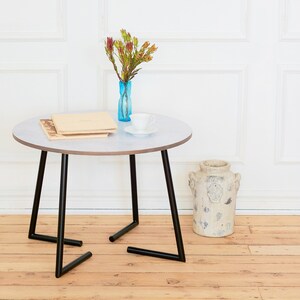May include: A round, white table with black metal legs. The table has two books and a white cup on top. A vase with brown flowers is on the table. The table is on a wooden floor.