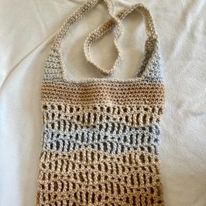 Könnte beinhalten: Eine gehäkelte Tasche mit einem langen Riemen. Die Tasche ist aus beige, hellbraunem und grauem Garn gefertigt. Die Tasche hat ein Netzdesign.