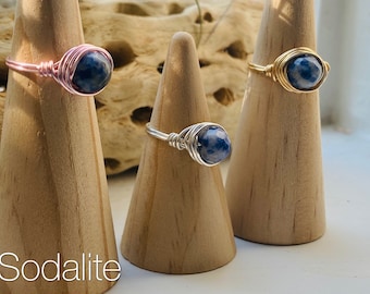 May include: Three wire-wrapped rings featuring faceted, round blue and white sodalite gemstones. Each ring is crafted with a different metal wire finish: one rose gold, one silver, and one gold. The word Sodalite is printed in white text.