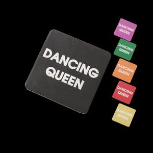 May include: A black square coaster with the words "DANCING QUEEN" in white. Several smaller square coasters in pink, green, orange, red, and yellow, each with the same text, are scattered around. All coasters are against a black background.