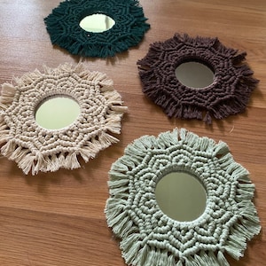 May include: Four small, round macrame mirrors in shades of green, brown, and beige. Each mirror has a circular reflective surface surrounded by intricate knotted macrame, with a fringed edge. The mirrors are arranged on a wooden surface.