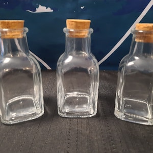 Puede incluir: Tres botellas de vidrio transparente con tapones de corcho. Las botellas son de forma cuadrada y tienen un fondo ligeramente redondeado.