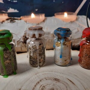 Könnte beinhalten: Vier kleine Glasgefäße mit Wachstropfen an der Außenseite. Jedes Gefäß enthält eine andersfarbige Mischung aus Kräutern und Kristallen. Die Gefäße sind in einer Reihe auf einer Holzoberfläche angeordnet.