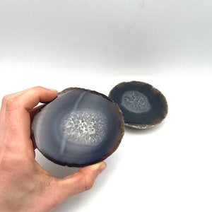 May include: Two polished agate slices. The agate has a dark blue-gray outer band, a lighter blue-gray inner band, and a white, crystalline center. The slices are on a white background.