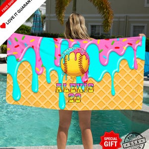 Könnte beinhalten: Ein Strandtuch im Softball-Design mit farbenfrohem Muster. Das Tuch zeigt einen gelben Softball mit einem rosa und blauen Donut-Design und den Namen "ALEXIS 23". Der Hintergrund hat ein Waffelmuster mit rosa und blau tropfendem Zuckerguss.