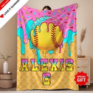 May include: Softball-themed blanket with a playful design. Features a yellow softball with red stitching, set against a pink background with sprinkles and dripping blue icing. The name "ALEXIS" and the number "6" are prominently displayed.