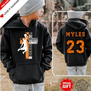 May include: Black hooded sweatshirt with a basketball graphic and the text "I can do all things through Christ who strengthens me." The back of the sweatshirt has the name "MYLES" and the number "23" in orange.