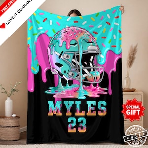 May include: A black blanket featuring a football helmet design with a donut theme. The helmet is teal with pink frosting and sprinkles. The name "MYLES" and the number "23" are displayed below the helmet in a glittery font. The blanket has the text "FREE SHIPPING" and "LOVE IT GUARANTEE".