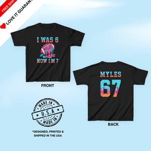 May include: Black t-shirt with a colorful football graphic that reads "I WAS 6 NOW I'M 7" on the front. The back of the shirt has the name "MYLES" and the number "67". The shirt is made in the USA.