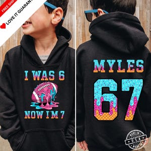 May include: Black hoodie with a colorful graphic. The front says "I WAS 6 NOW I'M 7" with a football design. The back has "MYLES" and the number "67" in a blue and pink design. Made in the USA.