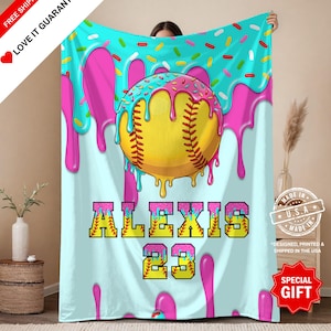 Könnte beinhalten: Eine hellblaue Decke mit einem Softball-Design. Sie zeigt einen gelb-rosa Softball, tropfendes rosa Zuckerguss und bunte Streusel. Der Name "ALEXIS" und die Nummer "23" sind abgebildet. Die Decke ist mit "MADE IN U.S.A." gekennzeichnet.