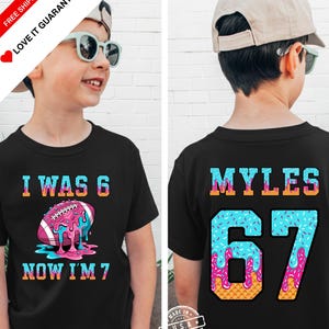 May include: Black t-shirt with a colorful football graphic and the text "I WAS 6 NOW I'M 7" on the front. The back of the shirt has the name "MYLES" and the number "67" in a similar colorful design. A fun birthday shirt.