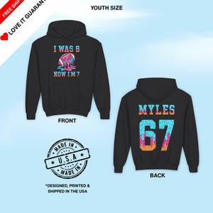 May include: Black youth-size hoodie with the text "I WAS 6 NOW I'M 7" on the front and "MYLES 67" on the back. The front design includes a football with colorful dripping accents. The back design features the name "MYLES" above the number 67.