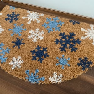May include: A half-circle doormat made of natural brown coir fiber. The mat features a pattern of snowflakes in white, light blue, and navy blue. The doormat is placed on a wooden floor.