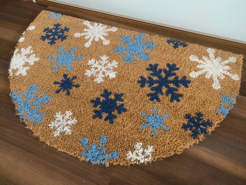 May include: A half-circle doormat made of natural brown coir fiber. The mat features a pattern of snowflakes in white, light blue, and navy blue. The doormat is placed on a wooden floor.