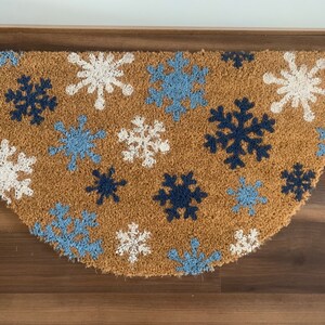 May include: A half-circle doormat made of natural coir fiber. The mat features a pattern of snowflakes in white, light blue, and navy blue against a brown background. The doormat is designed for indoor or outdoor use.
