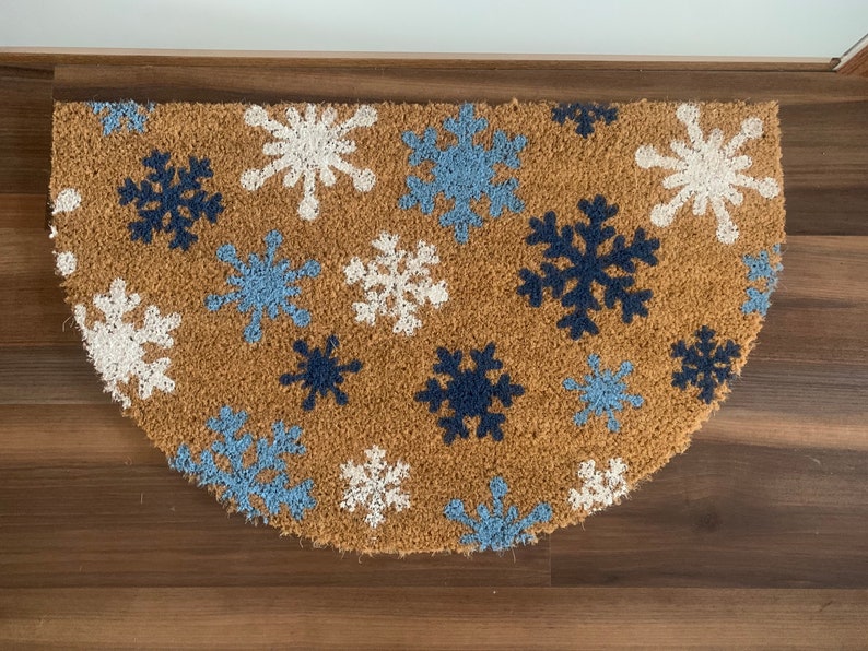 May include: A half-circle doormat made of natural coir fiber. The mat features a pattern of snowflakes in white, light blue, and navy blue against a brown background. The doormat is designed for indoor or outdoor use.