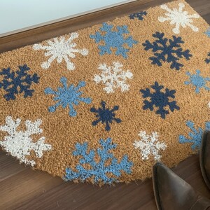 May include: A half-circle doormat made of natural coir fiber. The mat features a pattern of snowflakes in white, light blue, and navy blue. The mat is placed in front of a door, with a pair of brown boots visible.