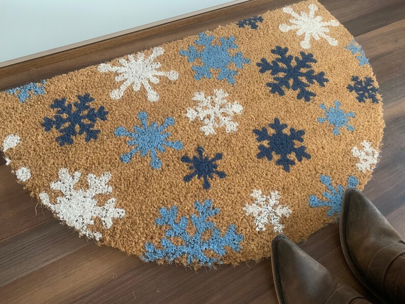 May include: A half-circle doormat made of natural coir fiber. The mat features a pattern of snowflakes in white, light blue, and navy blue. The mat is placed in front of a door, with a pair of brown boots visible.
