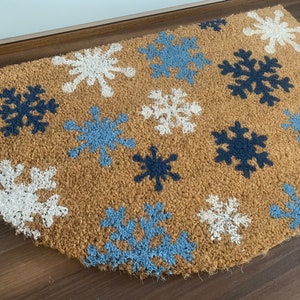 May include: A half-circle doormat made of natural brown coir fiber. The mat features a pattern of snowflakes in white, light blue, and navy blue. The snowflakes are evenly spaced across the surface of the mat.