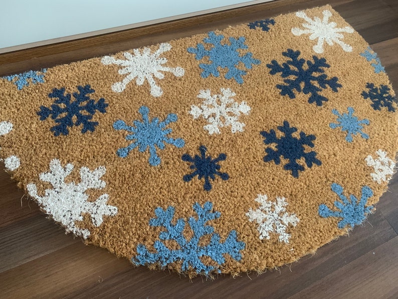 May include: A half-circle doormat made of natural brown coir fiber. The mat features a pattern of snowflakes in white, light blue, and navy blue. The snowflakes are evenly spaced across the surface of the mat.