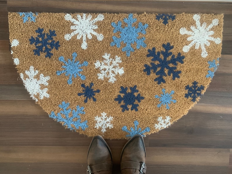 May include: A half-circle doormat made of natural coir fiber, decorated with snowflakes in white, light blue, and navy blue. The mat is placed on a wooden floor, with brown boots visible at the bottom.