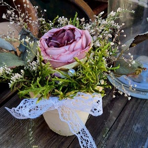 Könnte beinhalten: Ein kleiner, cremefarbener Topf gefüllt mit einer rosa-burgunderfarbenen Rose, umgeben von grünem Moos und zarten weißen Blüten. Ein weißes Spitzenband ist um den Topf gebunden. Die Anordnung steht auf einer verwitterten Holzoberfläche.