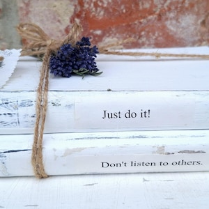 Könnte beinhalten: Zwei gestapelte Bücher mit weißer, abgenutzter Lackierung, zusammengebunden mit braunem Bindfaden. Auf dem oberen Buchrücken steht Just do it!, auf dem unteren Don't listen to others. Ein kleines Bündel getrockneter violetter Lavendel steckt unter dem Faden.