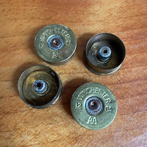 Peut inclure: Quatre douilles de cartouches de fusil de chasse Winchester 12 GA en laiton. Deux douilles sont intactes et deux sont coupées, révélant les composants internes. Les douilles sont sur une surface en bois.