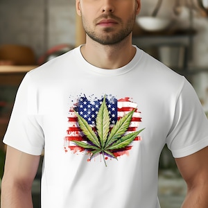 Puede incluir: Una camiseta blanca con una hoja de marihuana verde impresa en ella. La hoja está superpuesta a una bandera estadounidense de estilo acuarela.