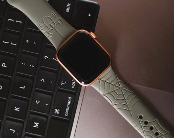 May include: A gray Apple Watch with a spiderweb patterned band on a black laptop keyboard. The watch face is black and the band is made of silicone.