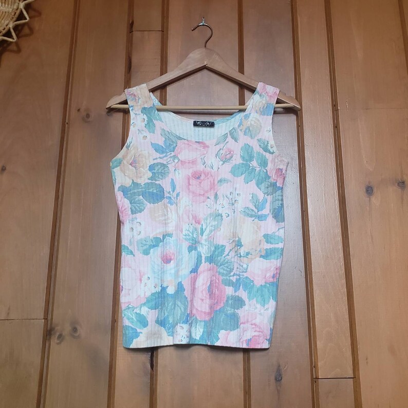 May include: A sleeveless, pink and white floral print tank top with a ribbed texture. The tank top has a scoop neck and is hanging on a wooden hanger.