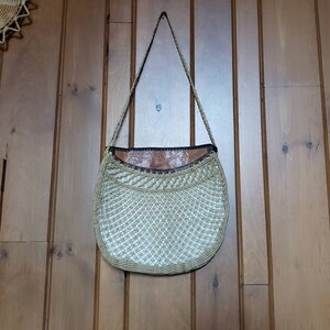 May include: A beige crocheted bag with a brown leather flap and a long strap. The bag is hanging on a wooden wall.
