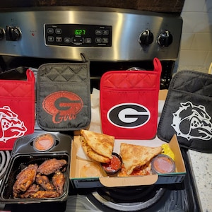 May include: Four red and black oven mitts with a white bulldog graphic and a white 'G' graphic. The mitts are on a counter next to a box of pizza and chicken wings.