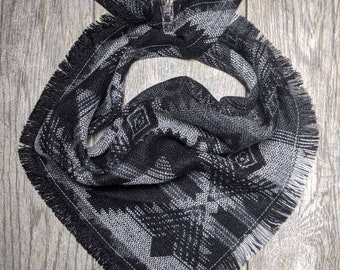May include: A black and gray patterned bandana with a fringed edge. The bandana features a geometric design with diamond and arrow-like shapes. The fabric is folded and tied at the top, creating a stylish accessory.
