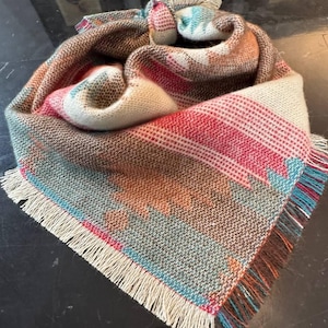 May include: A multi-colored woven scarf with fringe. The scarf is made of a soft, warm material and features a geometric pattern in shades of brown, red, blue, and white.
