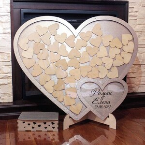 Puede incluir: Un libro de visitas de madera en forma de corazón plateado con un marco en forma de corazón más pequeño en el interior. El marco más pequeño tiene el texto "Roman & Elena 21.08.2021" escrito en negro. El corazón más grande está lleno de pequeños corazones de madera.