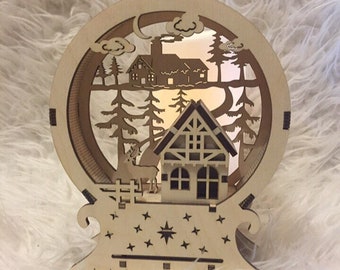 Può includere: Una scena di villaggio natalizio in legno con una casa, alberi e un cervo. La scena è illuminata da una luce calda. La scena è circondata da una cornice circolare con una luna e stelle.